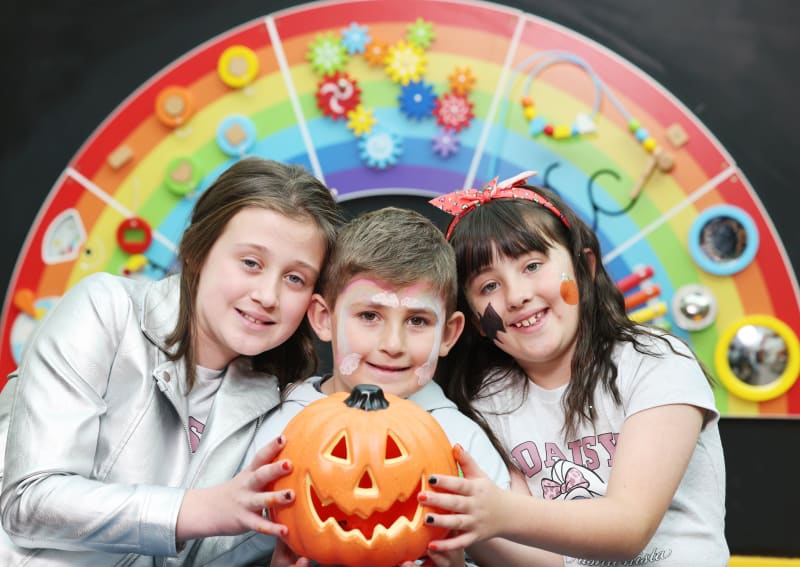 Three children with face paint smile while holding a carved jack-o'-lantern in front of a rainbow sensory play wall.