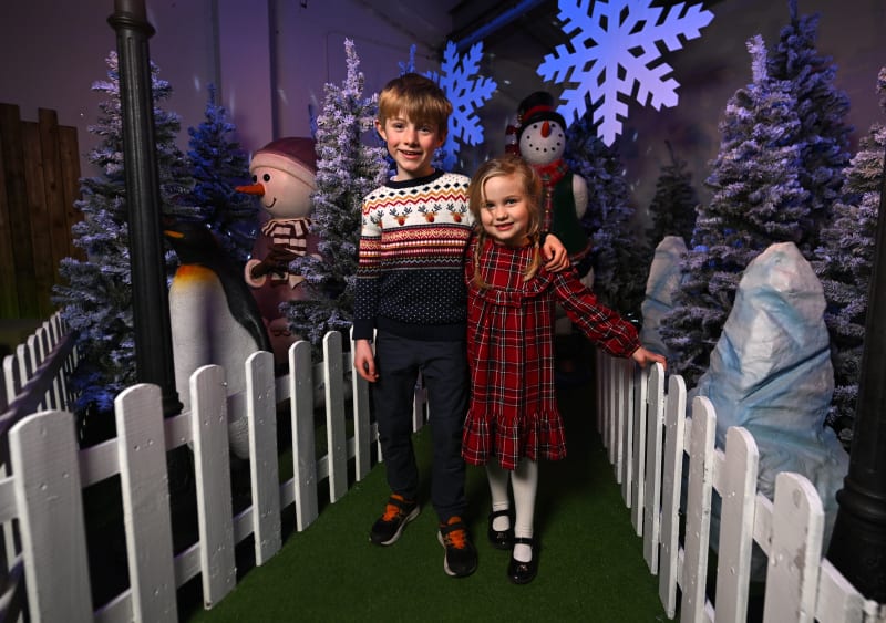 Two children pose in a Christmas winter wonderland grotto with snowmen, trees, and snowflake decorations.