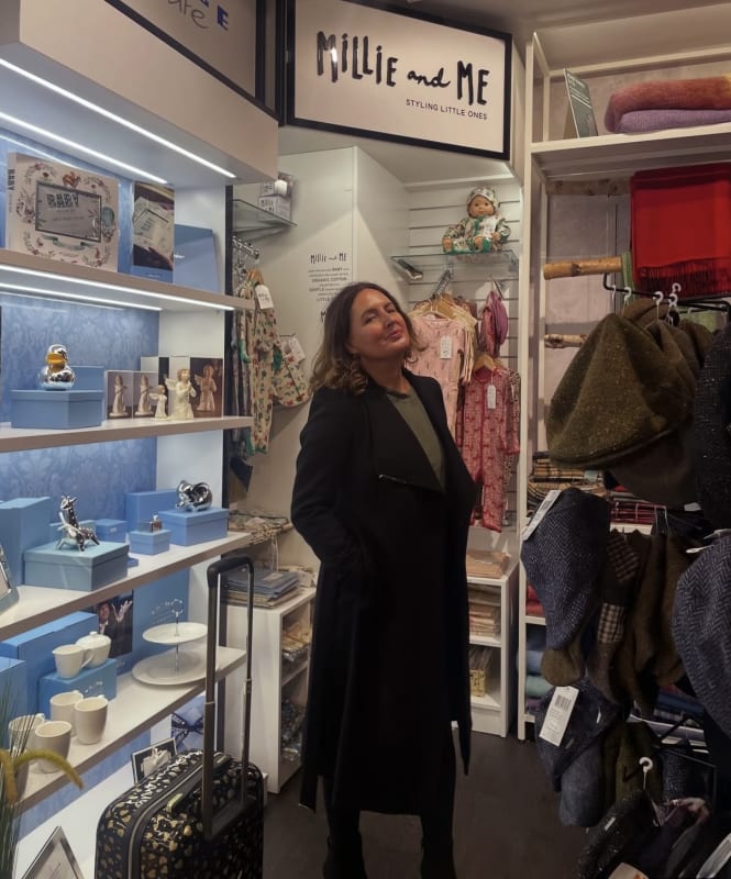 Woman in black coat standing in front of Millie and Me children's boutique display with gifts and clothing.