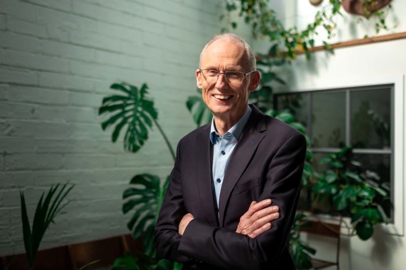 An Tollamh Pádraig Ó Duibhir smiling with arms crossed, wearing a dark blazer and light blue shirt, indoors with green plants.