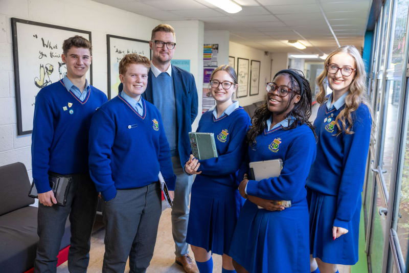 Five secondary school students in blue uniforms smile with an adult male teacher in a school corridor.