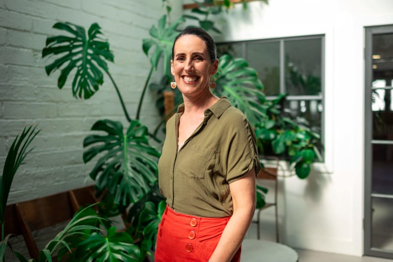 Dr Cáit Verling smiling indoors, wearing an olive shirt and red skirt, with monstera plants behind her.